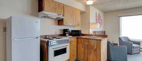 A compact kitchen with wooden cabinets, a white refrigerator, a stove, and a microwave. There is a sink and two chairs in the adjacent living area.