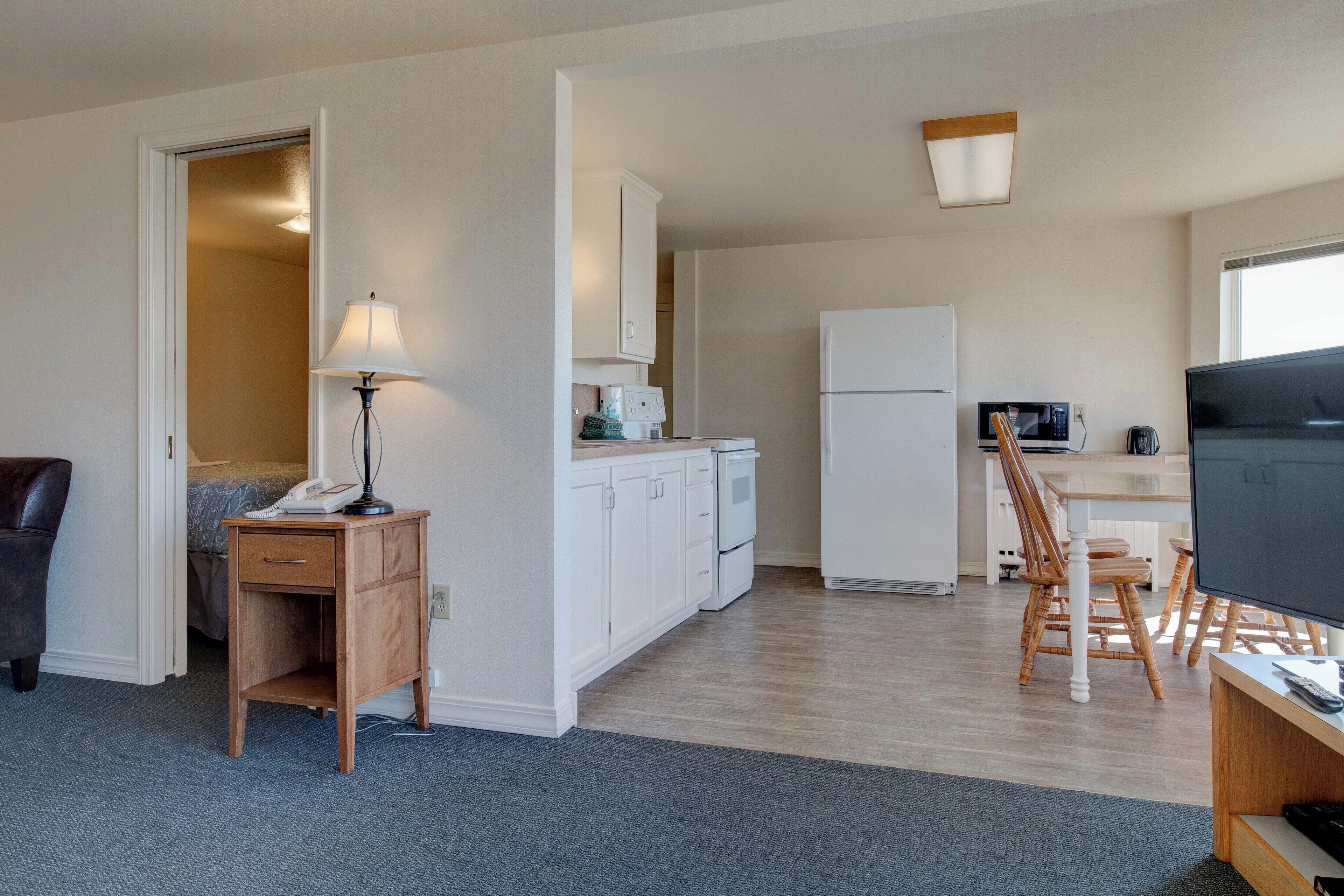 Un espacio habitable compacto con kitchenette, una zona de comedor con sillas de madera, un escritorio pequeño con lámpara y un televisor.