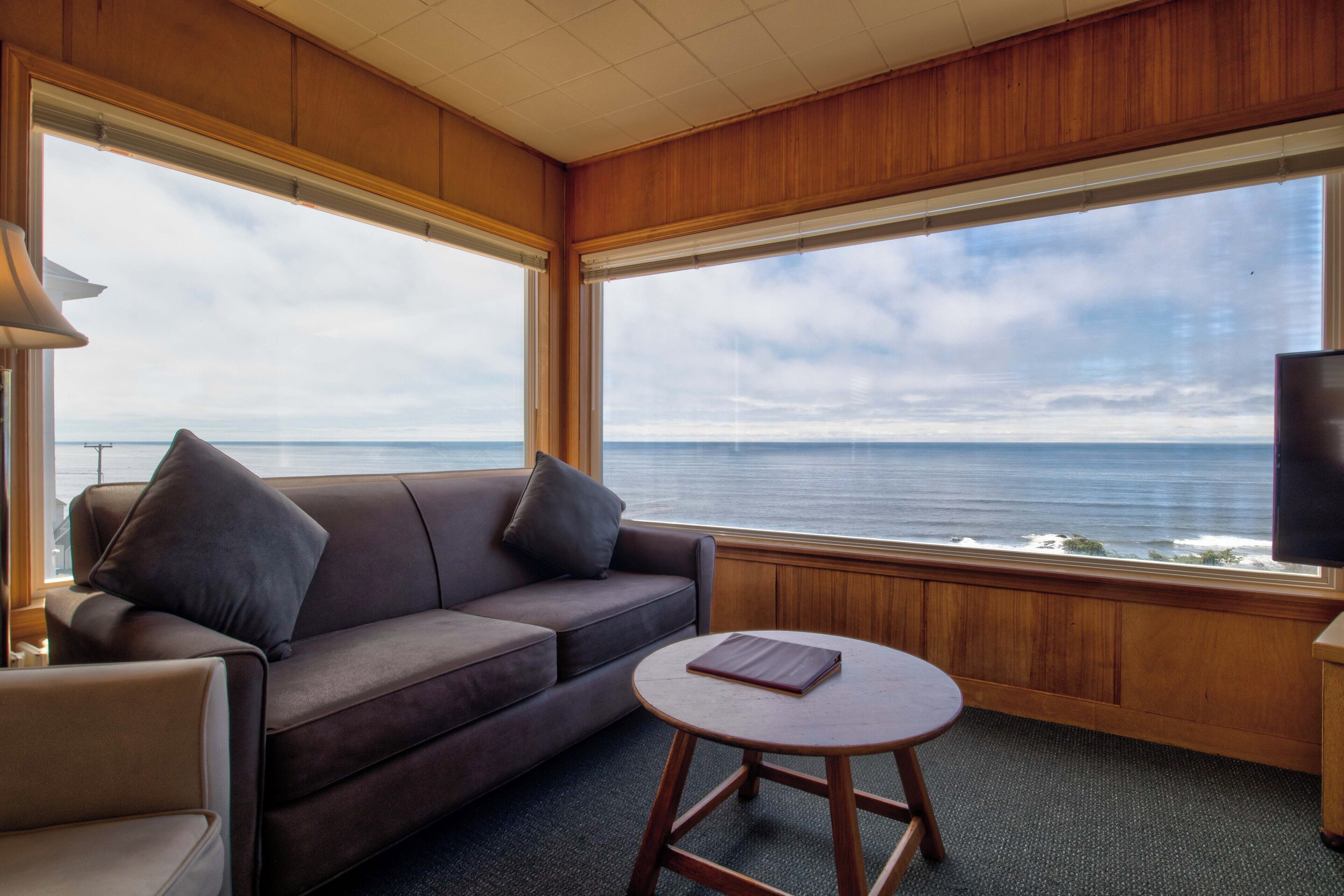 Ein Zimmer mit einer braunen Ledersofa, einem kleinen runden Holztisch und einem großen Fenster mit Blick auf den Ozean.
