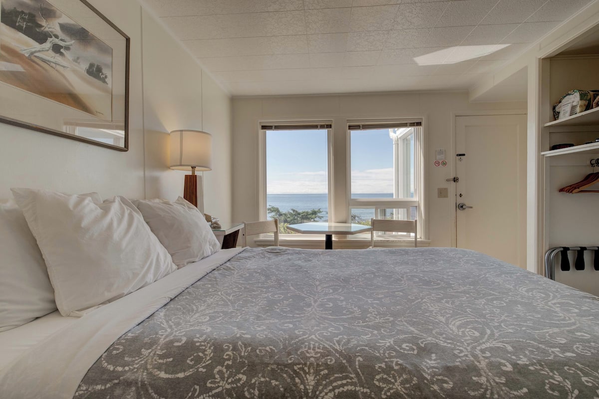 A bedroom with a large bed, a view of the sea through a door, and a small table and chair.