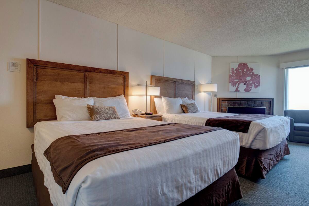 A hotel room with two beds, each with a wooden headboard, white bedding, and a brown throw blanket. There is a flat-screen TV mounted on the wall, a lamp on each nightstand, and a painting above the TV.