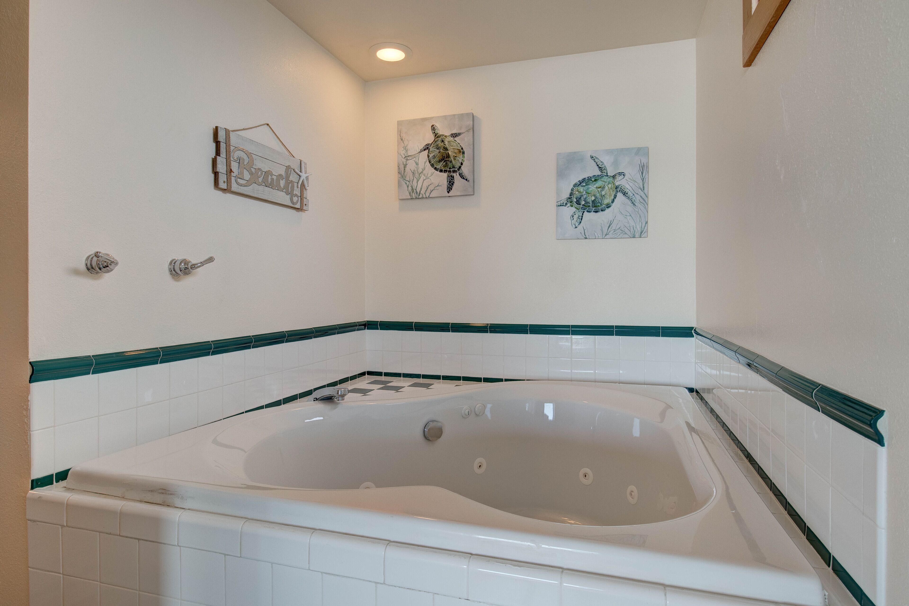 A bathroom with a large bathtub, white tiles with green borders, and three wall-mounted artworks depicting sea turtles.