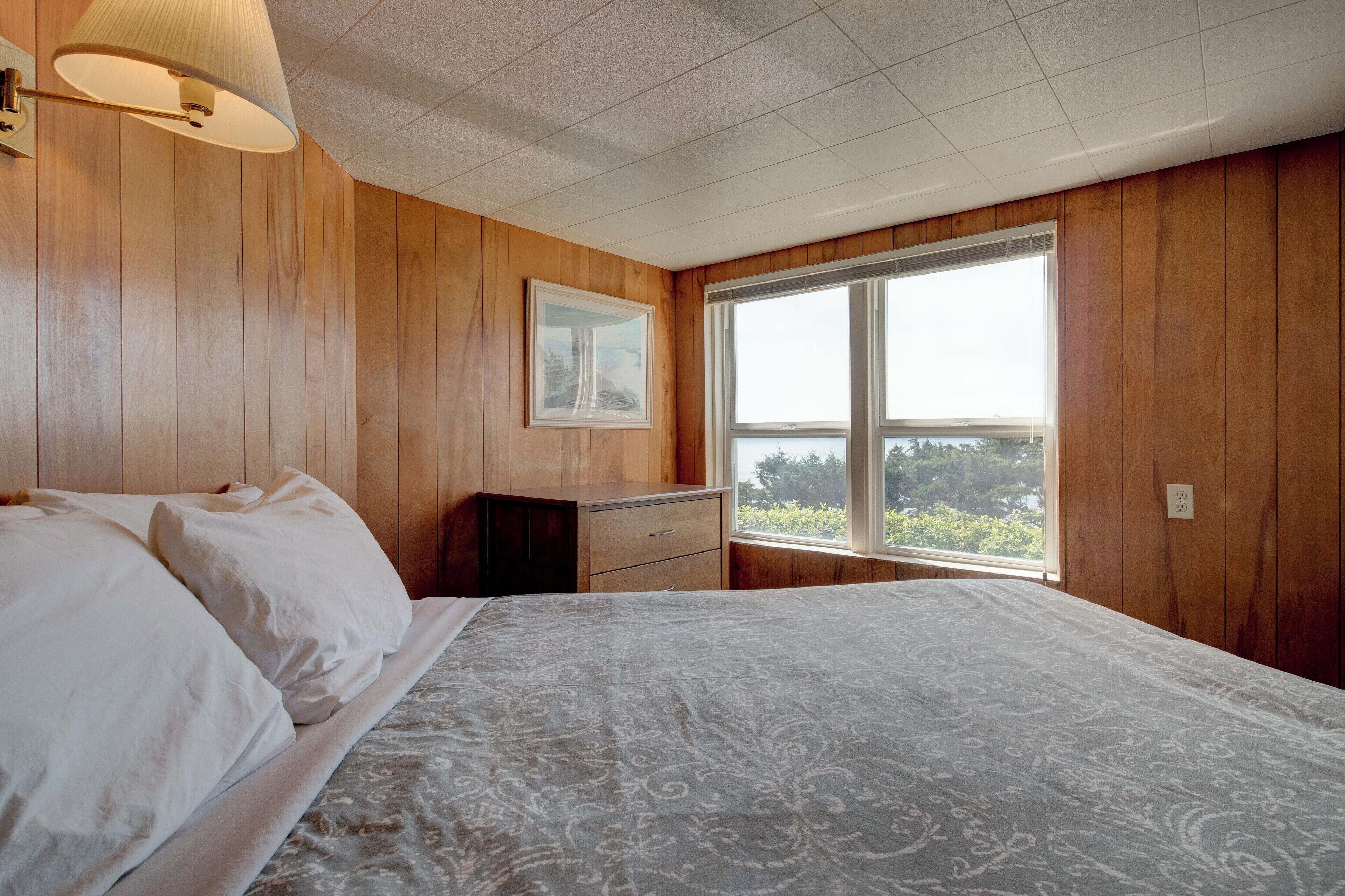 Een slaapkamer met houten lambrisering, een bed met witte lakens, een nachtkastje, een kaptafel en een raam met uitzicht op groen.