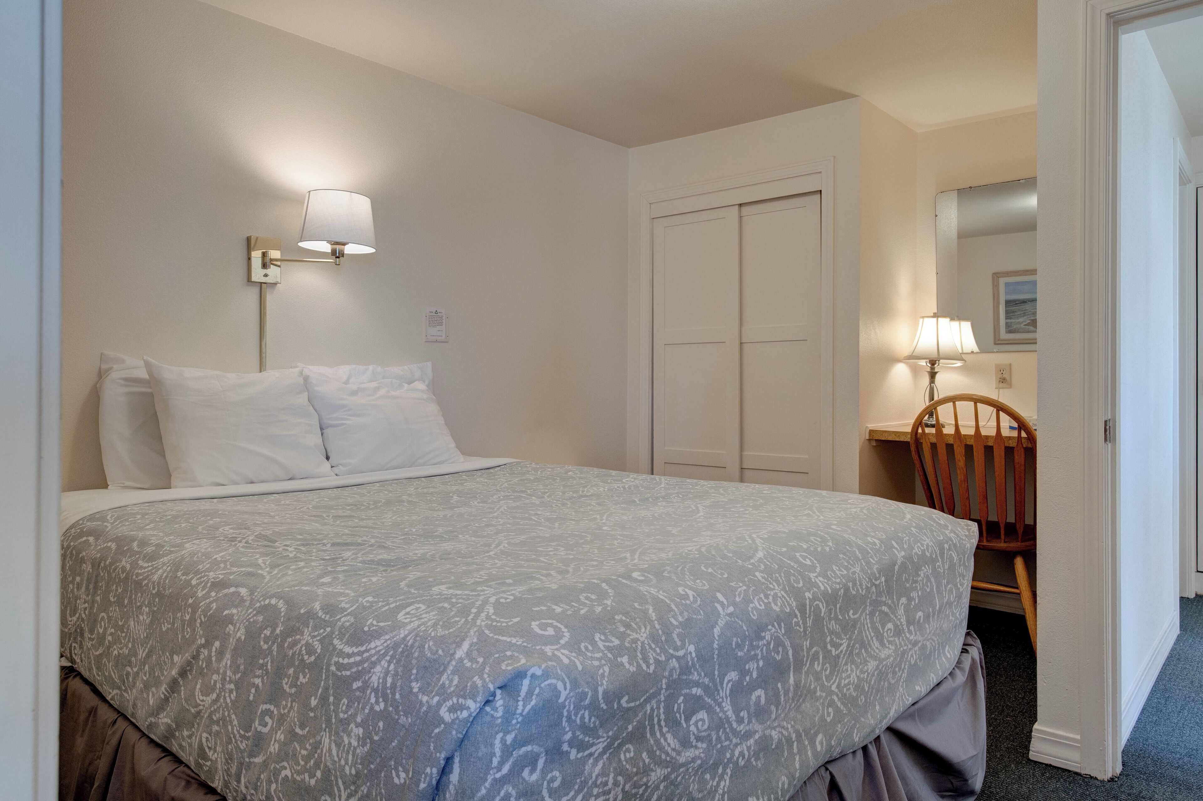 A neatly made bed with a patterned bedspread, a bedside lamp, a wooden chair, and a closed door in a hotel room.