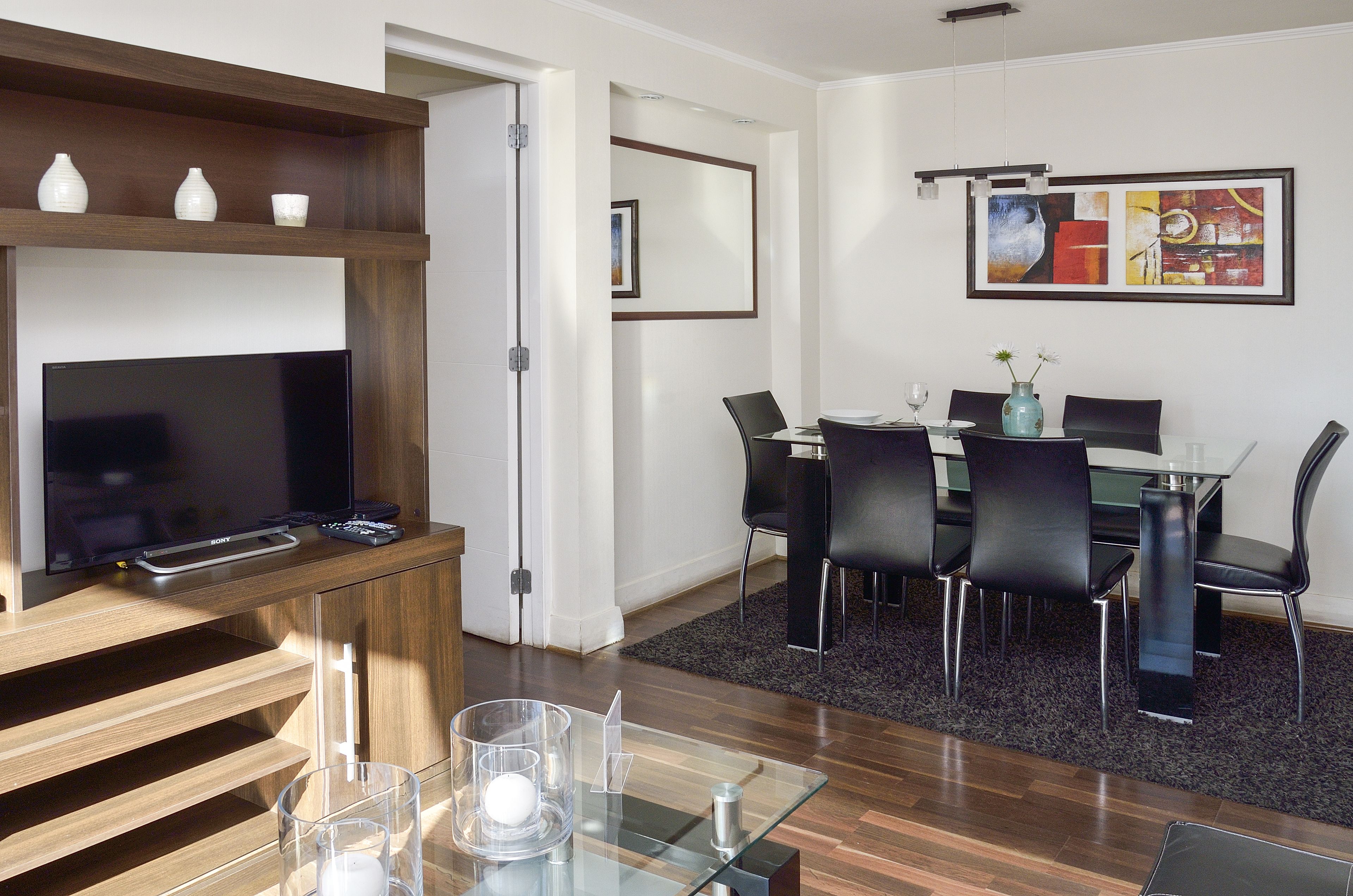 Una sala moderna con un televisor de pantalla plana, estanterías de madera, una mesa de centro de vidrio y un comedor con una mesa de vidrio y sillas negras.