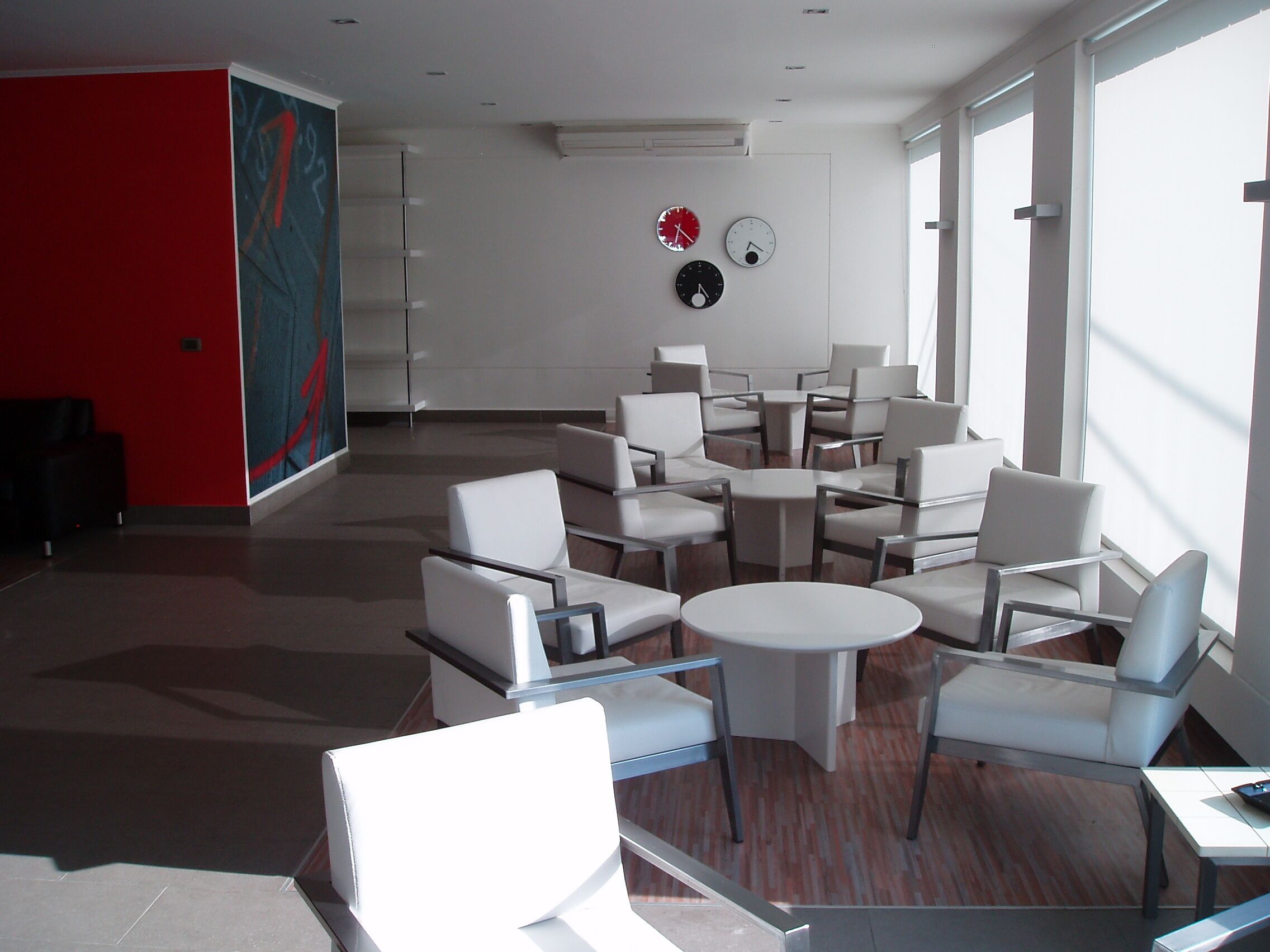 A modern lounge area with white chairs and tables, a red accent wall, and clocks on the wall.