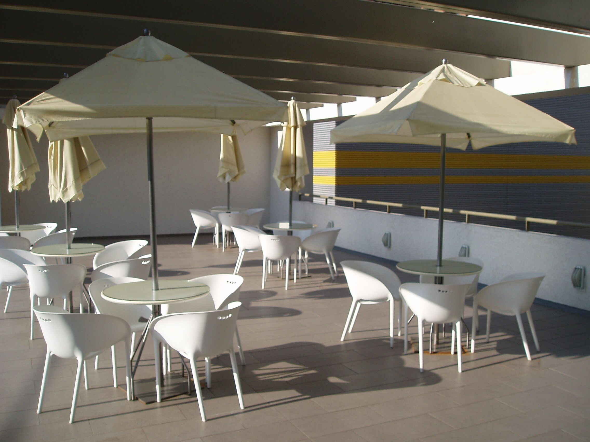 An outdoor patio area with white tables and chairs, beige umbrellas, and a striped wall in the background.