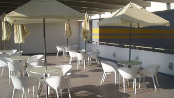 An outdoor patio area with white tables and chairs, beige umbrellas, and a striped wall in the background.