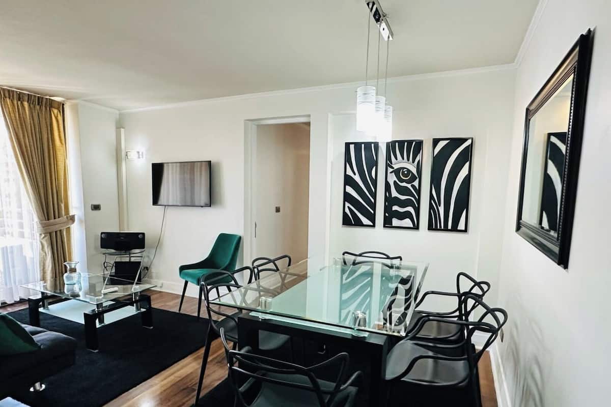 Una sala de estar moderna con una mesa de comedor de vidrio, sillas negras, un sillón verde azulado y un televisor de pantalla plana, además de arte abstracto en la pared.