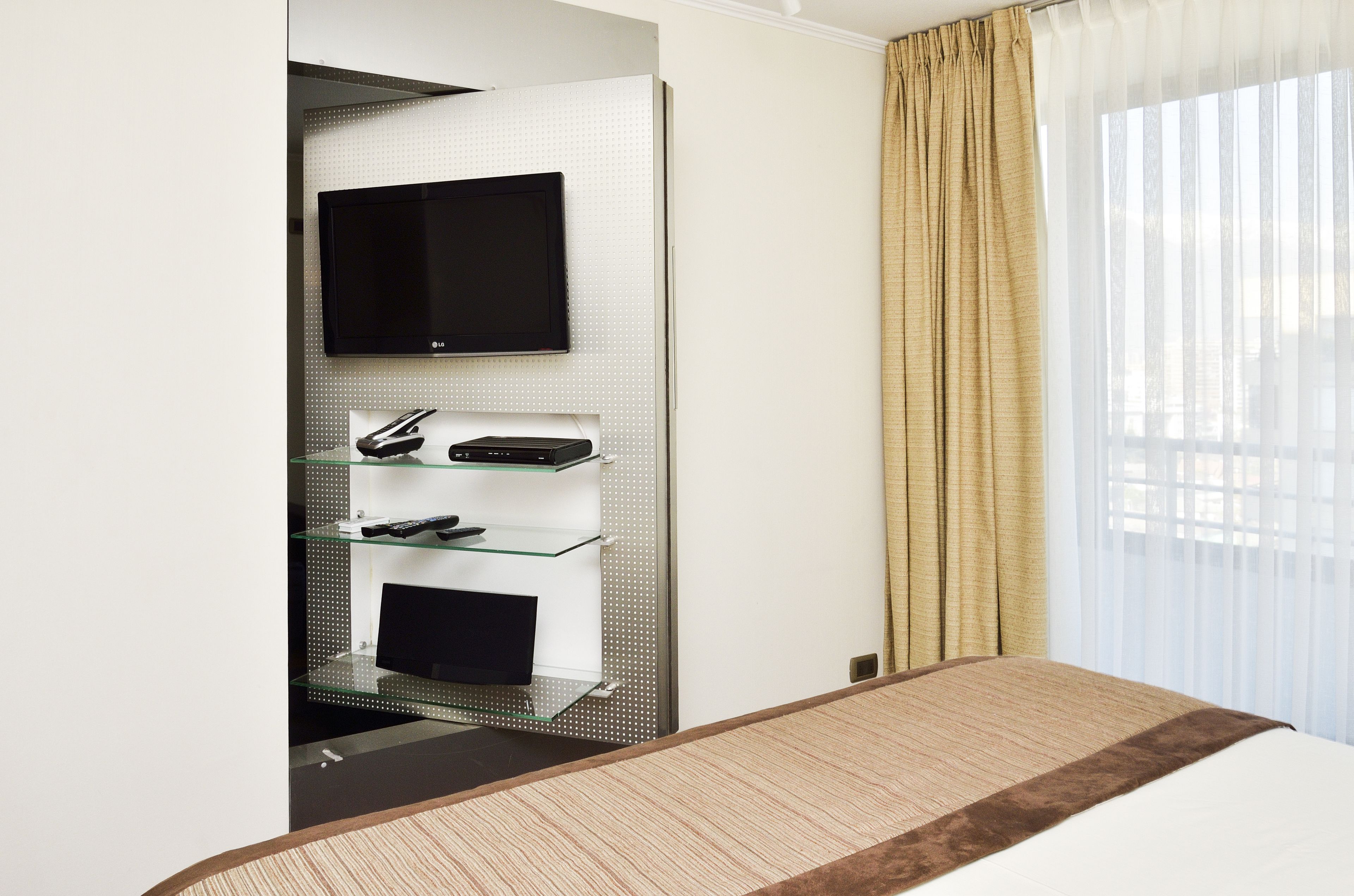 Une chambre d’hôtel équipée d’un lit, d’un téléviseur à écran plat fixé au mur et d’une étagère en verre contenant des appareils électroniques.