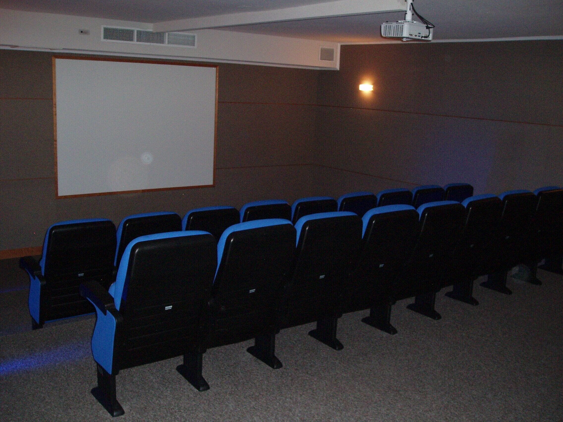 Une salle de cinéma avec des rangées de sièges noirs et bleus, un écran blanc et un projecteur fixé au plafond.