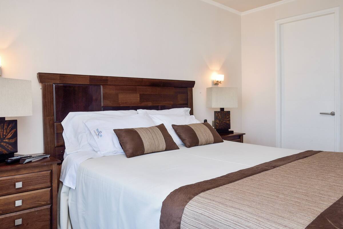 Una cama bien hecha con ropa de cama blanca y marrón, cabecera de madera, mesitas de noche con lámparas y una puerta blanca.