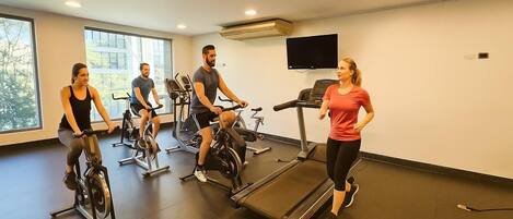Un gimnasio con cuatro personas usando equipo de cardio, un televisor en la pared y amplias ventanas.