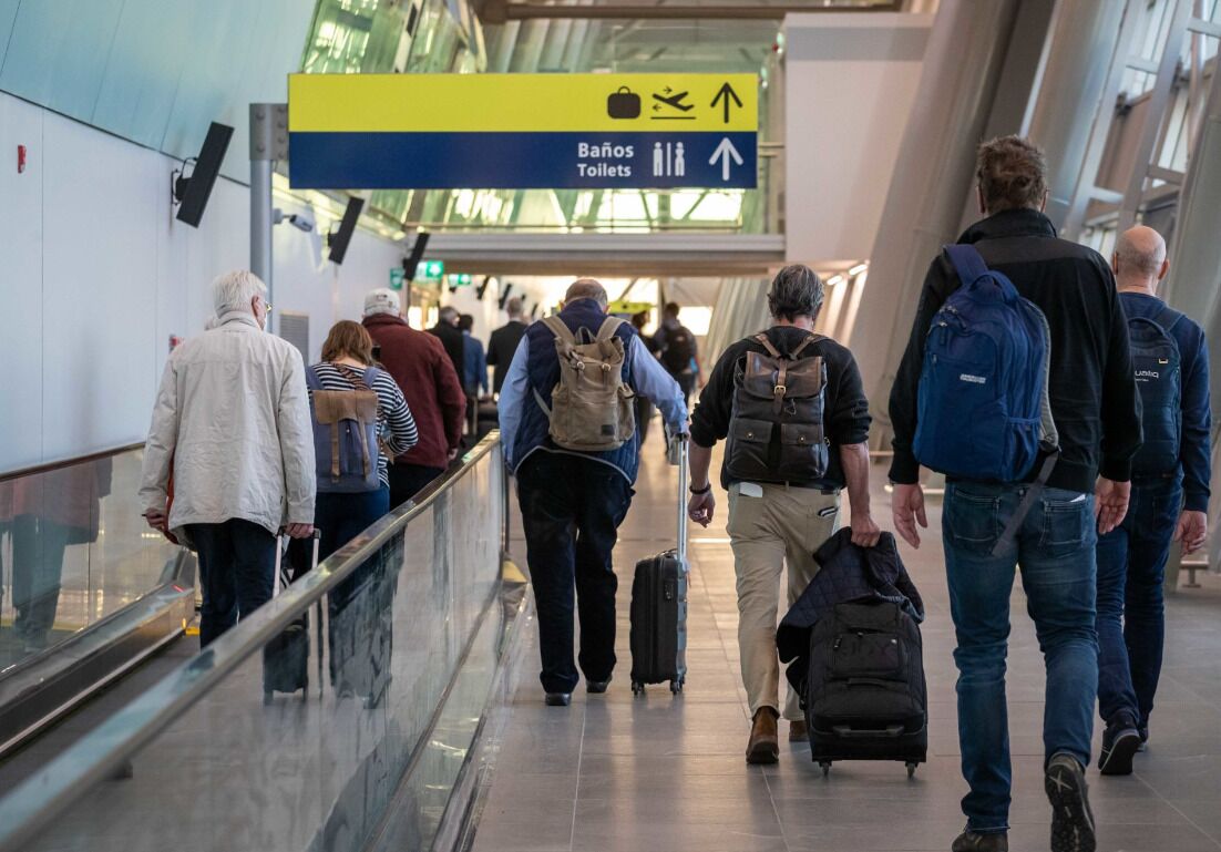 Een drukke corridor op de luchthaven, met reizigers die naar het terminalgebouw lopen, een bord met da op 'toiletten' en een bagagewagen.