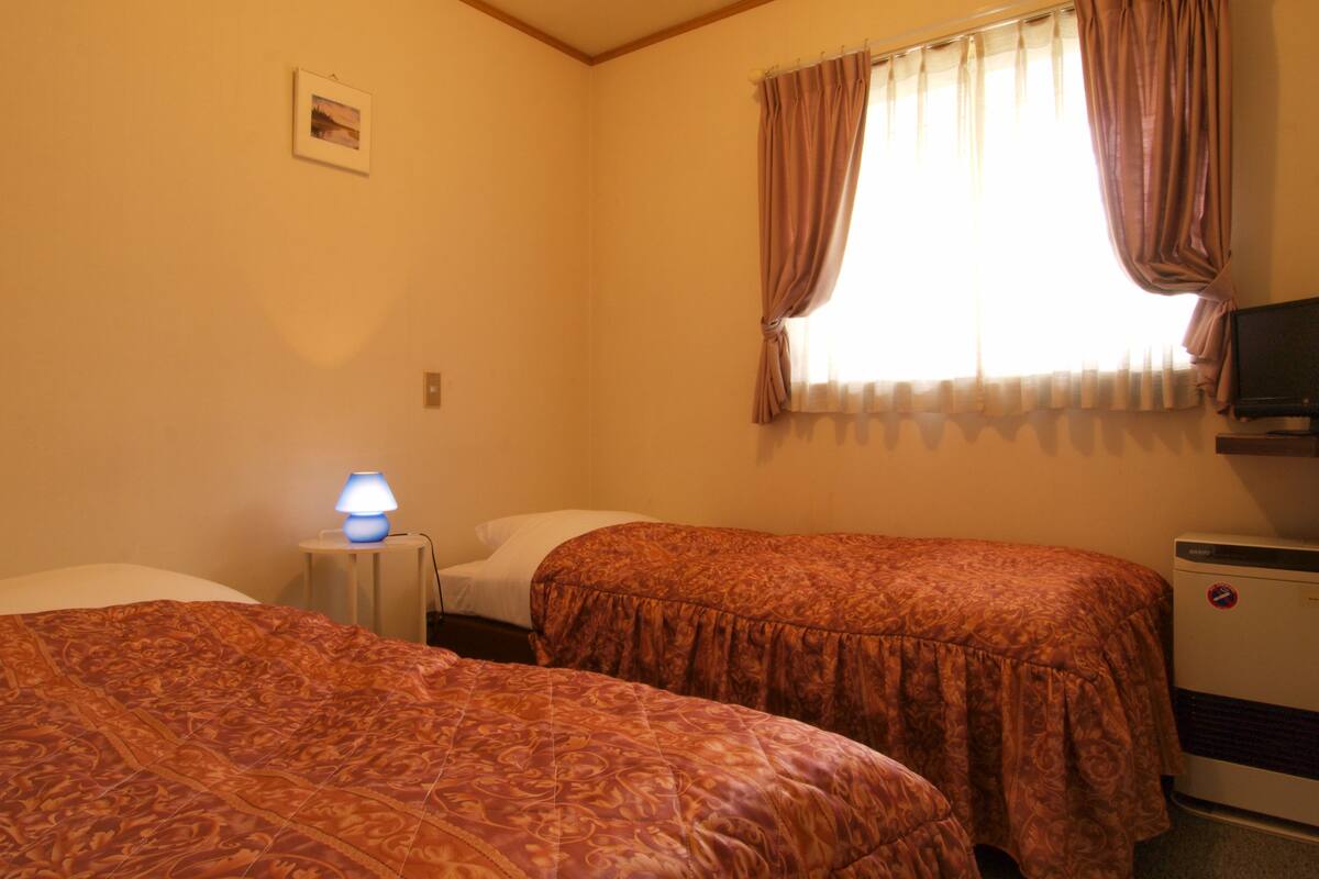 A room with two beds, a small table with a lamp, a TV, and a window with curtains.