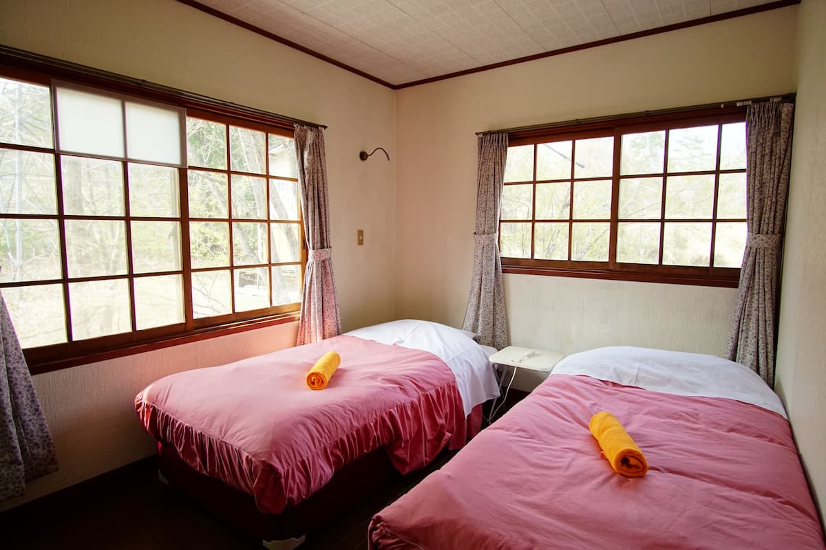 A room with two beds, each with a pink bedspread and a rolled yellow towel. There are large windows with curtains, and a small table between the beds.