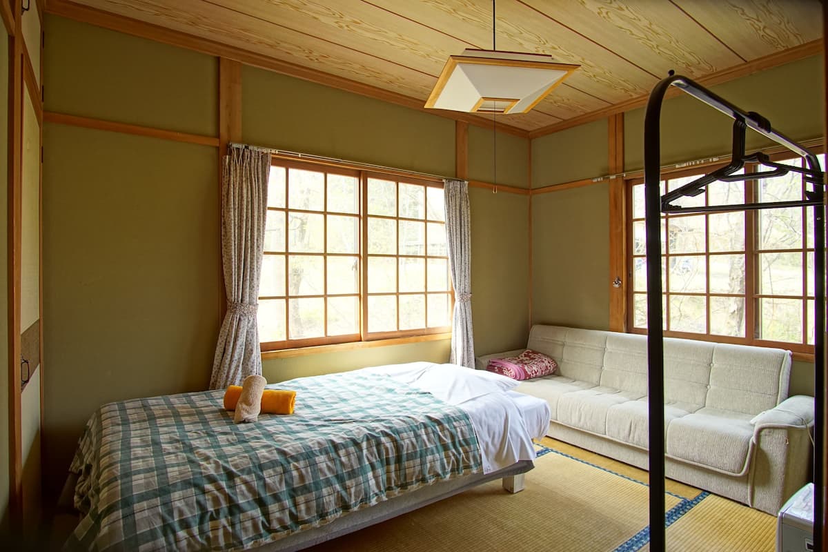 A traditional Japanese-style room with tatami flooring, a futon bed, a low wooden ceiling, and large windows with curtains.