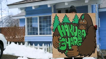 A person holding a sign with a cartoon bear and trees, in front of a blue house with a white picket fence.