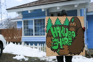 A person holding a sign with a cartoon bear and trees, in front of a blue house with a white picket fence. - share&guesthouse HAKUBA SHARE - Hostel (Hakuba)