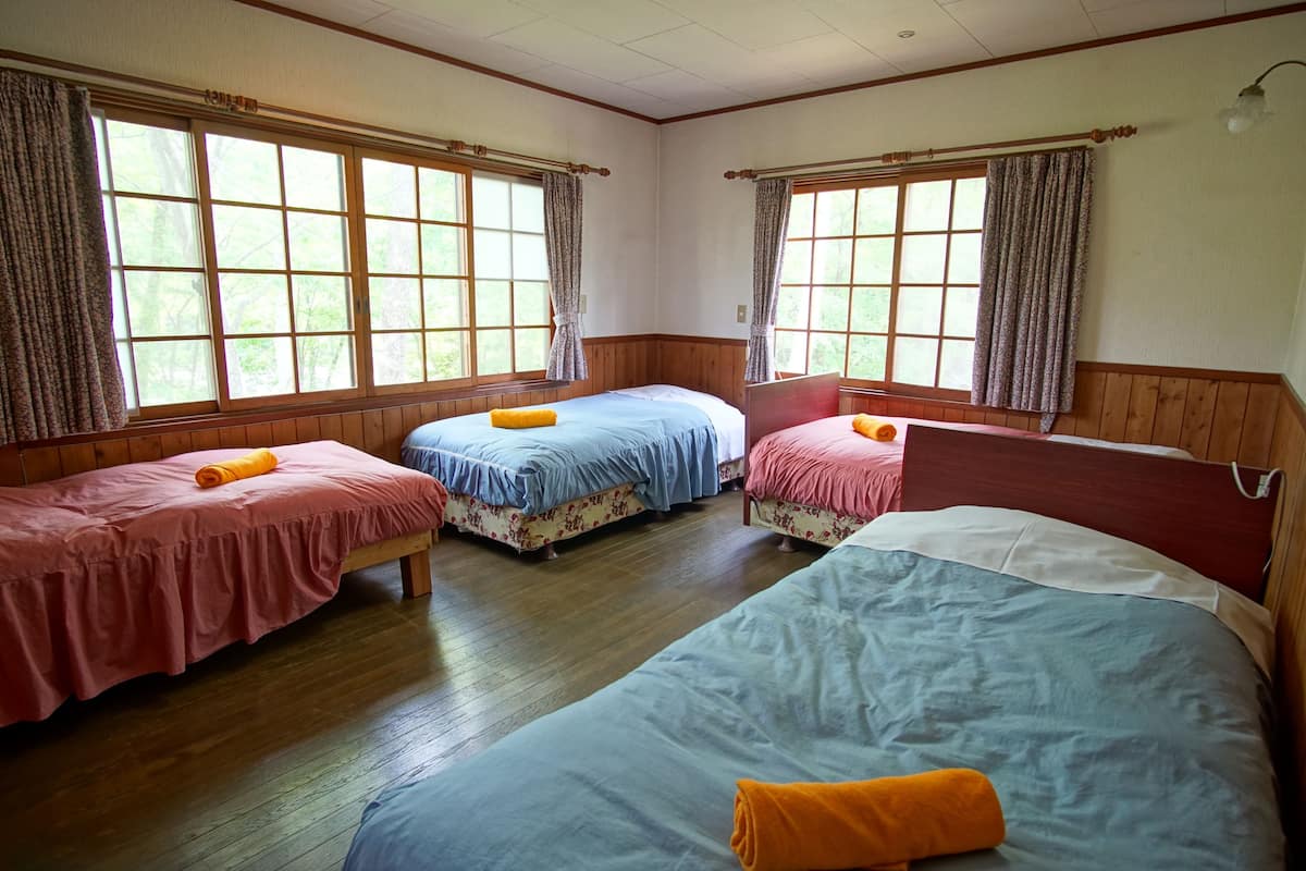 A room with three beds, wooden floors, and large windows with curtains.
