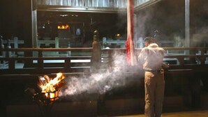Eine Person steht an einem Grill mit sichtbaren Flammen, draußen vor einem Holenzaun und einem Gebäude im Hintergrund.