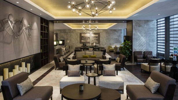 A modern hotel lobby with a central seating area, a chandelier, and a view of the kitchen in the background. - North star Hangzhou Expo Center Hotel (Hangzhou)
