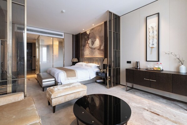 Una habitación de hotel moderna con una cama grande, un baño con paredes de vidrio y un armario de tonos oscuros, además de una mesa redonda negra.