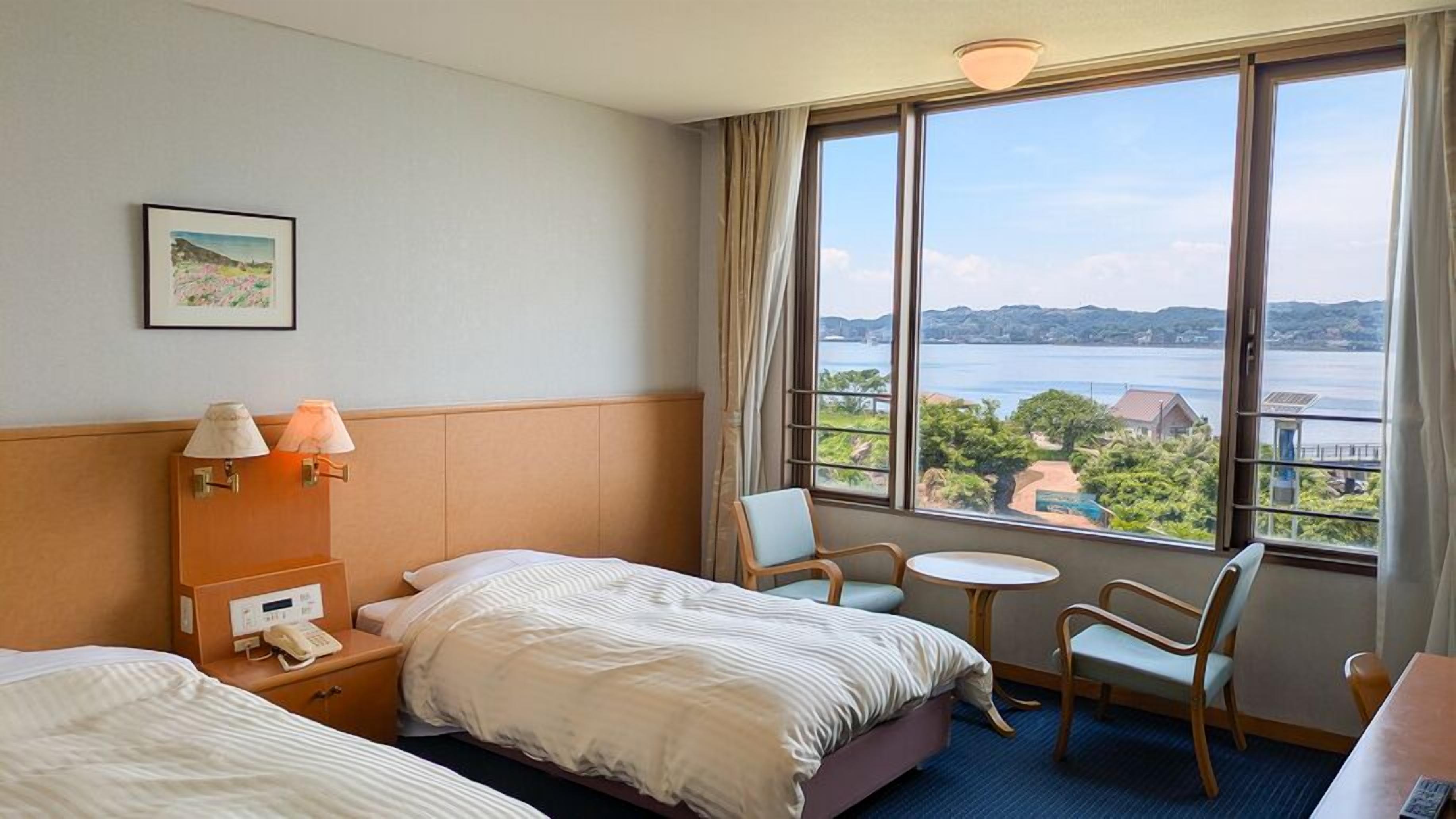 Ein Hotelzimmer mit zwei Betten, einem kleinen Tisch, einem Stuhl, einer Lampe, einem Telefon und einem großen Fenster mit Blick auf ein Gewässer und Gebäude.