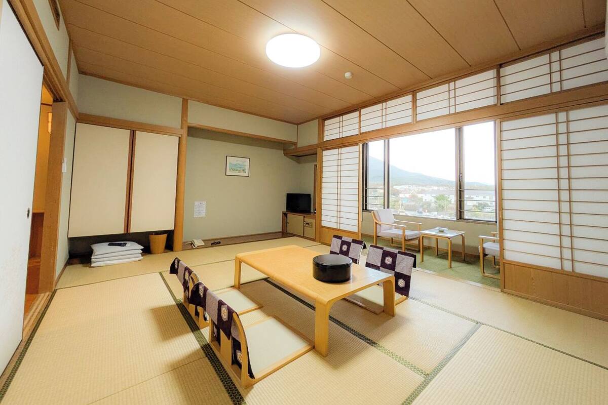 Une pièce de style japonais traditionnel, avec un sol recouvert de tatamis, une petite table basse entourée de coussins, un téléviseur et une vue sur une montagne, visible à travers de grandes fenêtres.