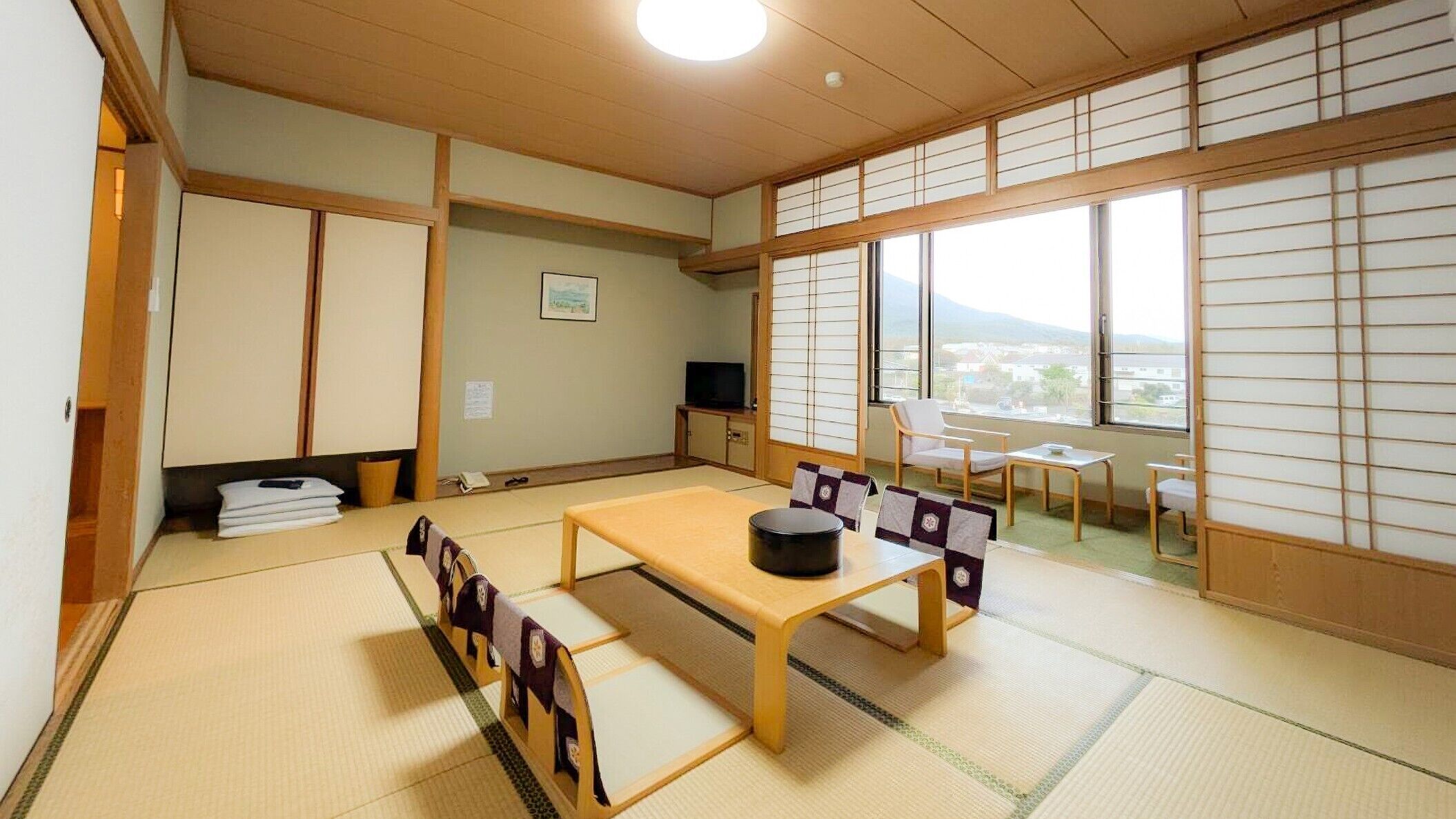 Ein traditioneller japanischer Raum mit Tatami-Böden, einem niedrigen Tisch mit Kissen, Schiebetüren und Blick durch große Fenster.