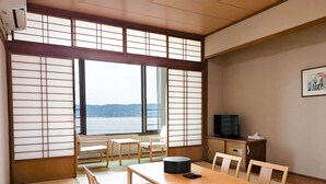 A traditional Japanese room with tatami flooring, a low table with cushions, a television on a cabinet, and a view of a body of water through shoji screens. - Kokuminshukusha Rainbow Sakurajima (Kagoshima)
