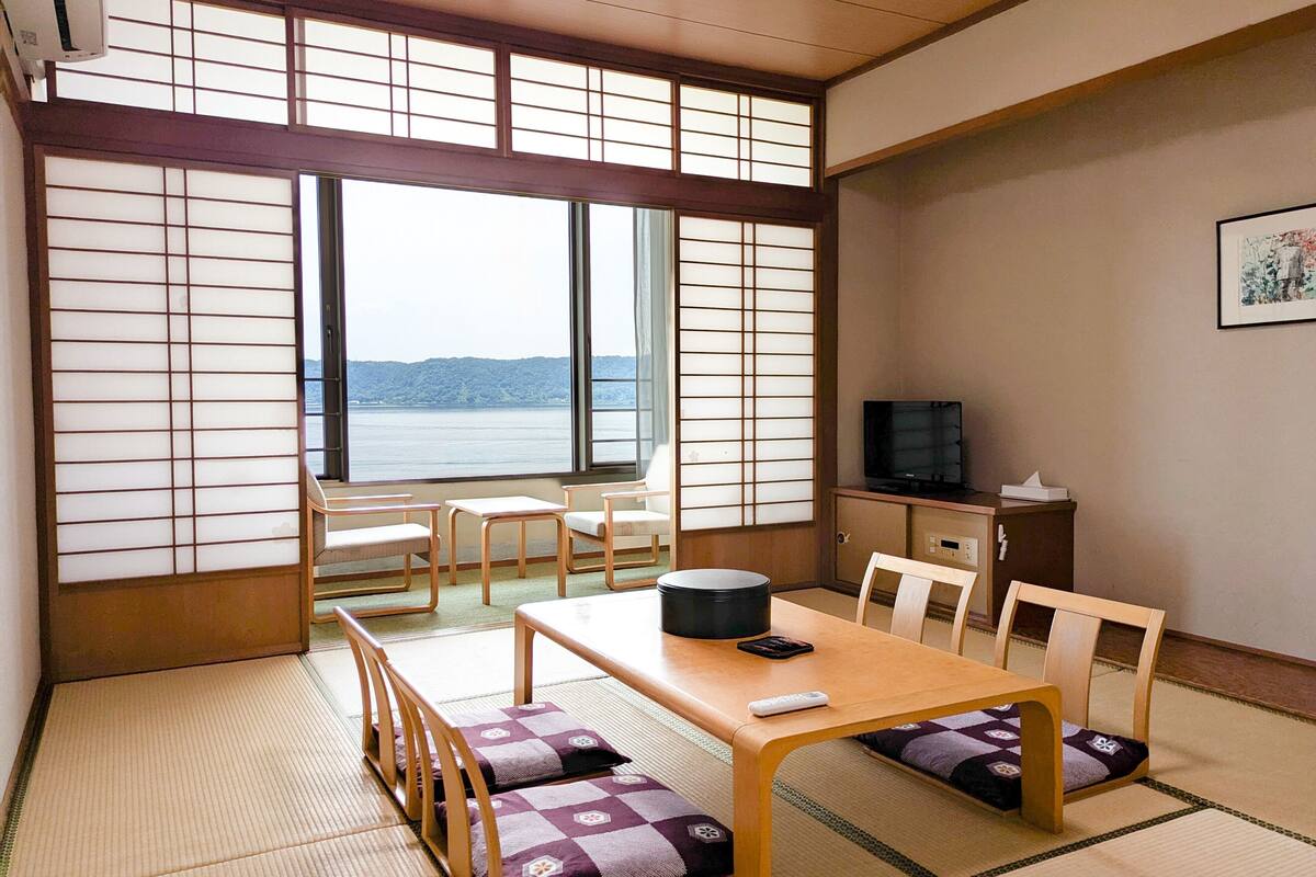Une pièce de style japonais traditionnel, avec un sol recouvert de tatamis, une table basse avec des coussins, un téléviseur posé sur un meuble et une vue sur un plan d’eau à travers des panneaux coulissants.
