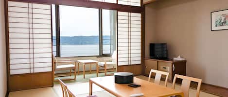 A traditional Japanese room with tatami flooring, a low table with cushions, a television on a cabinet, and a view of a body of water through shoji screens.