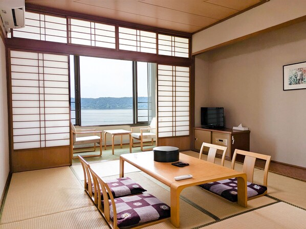 A traditional Japanese room with tatami flooring, a low table with cushions, a television on a cabinet, and a view of a body of water through shoji screens.