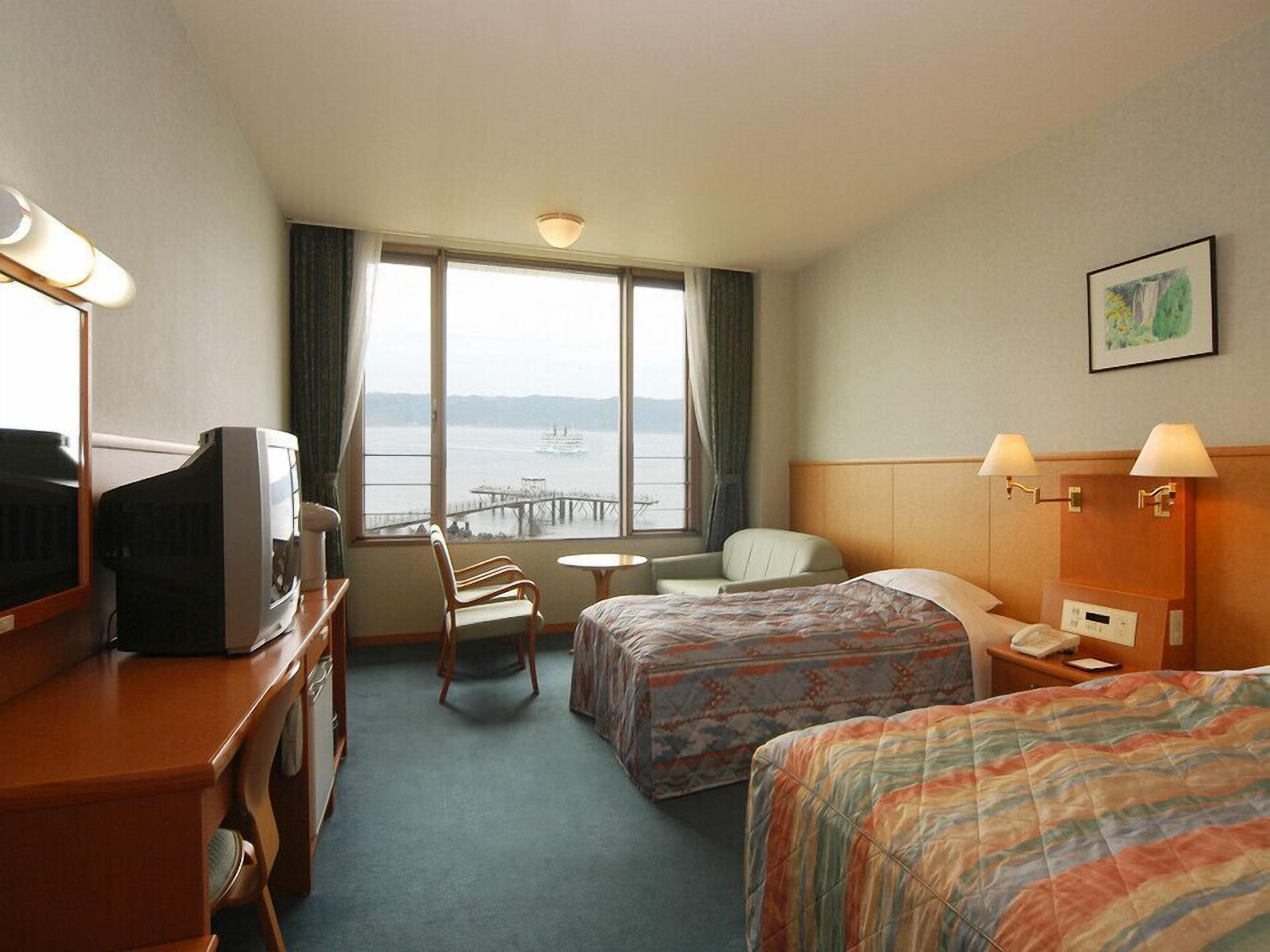 Ein Hotelzimmer mit zwei Betten, einem Schreibtisch mit Fernseher, einem Sessel, einem kleinen Tisch und Blick aufs Wasser.
