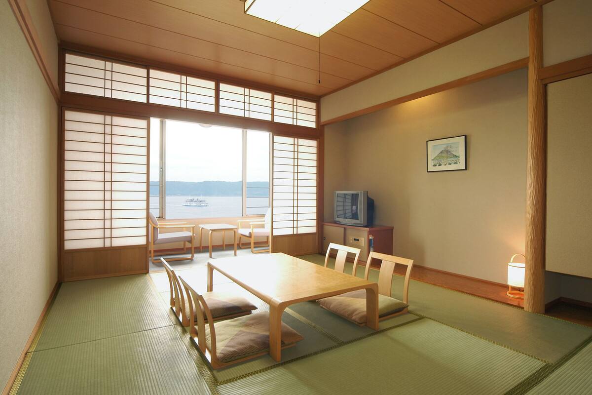Une pièce de style japonais traditionnel, avec un sol recouvert de tatamis, une petite table basse entourée de coussins et un téléviseur posé sur un meuble en bois.