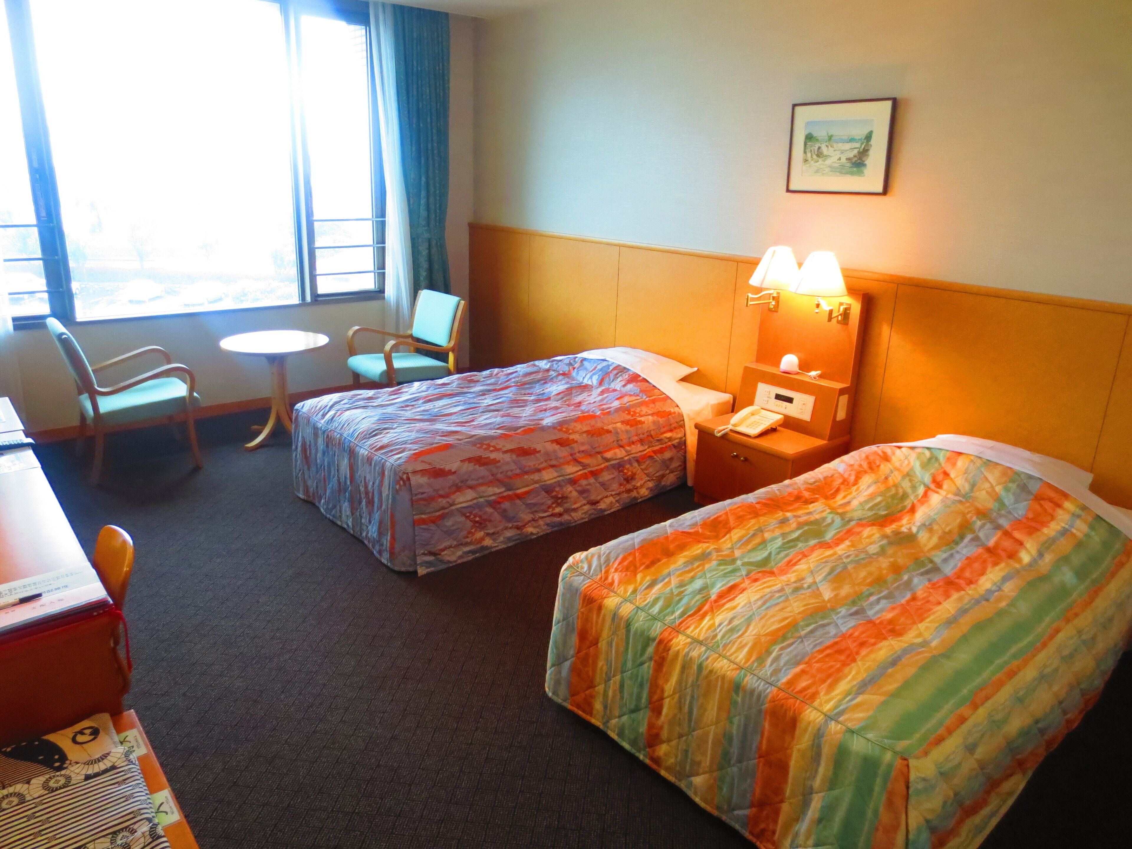 Ein Hotelzimmer mit zwei Betten, einem Schreibtisch, einem Stuhl, einem kleinen Tisch und einem Fenster mit Vorhängen.