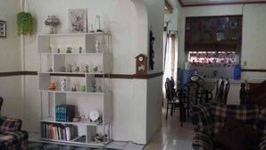 A living room with a white bookshelf, a dining area with a table and chairs, and a kitchen area with cabinets and a window. - Kensington Garden BNB (Baguio)