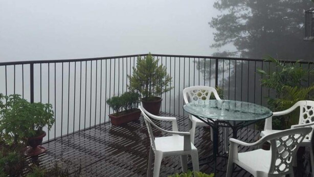 A balcony with a glass table and white chairs, surrounded by potted plants and overlooking a body of water. - Kensington Garden BNB (Baguio)