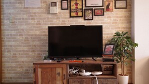 A brick wall with framed pictures, a television on a wooden stand, and a potted plant. - Kokoro House (Osaka)