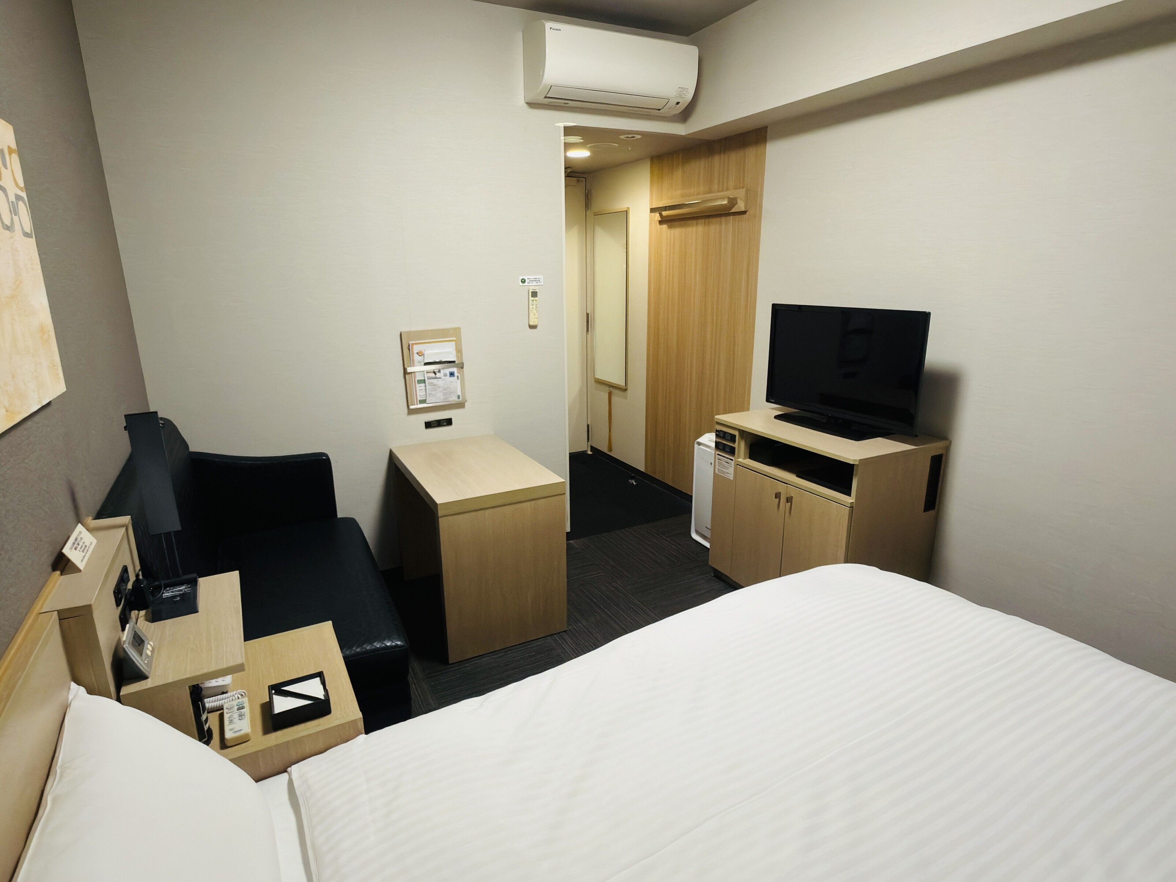 Habitación de hotel con cama, escritorio con silla, televisor sobre un mueble de madera, refrigerador pequeño y aire acondicionado de pared.