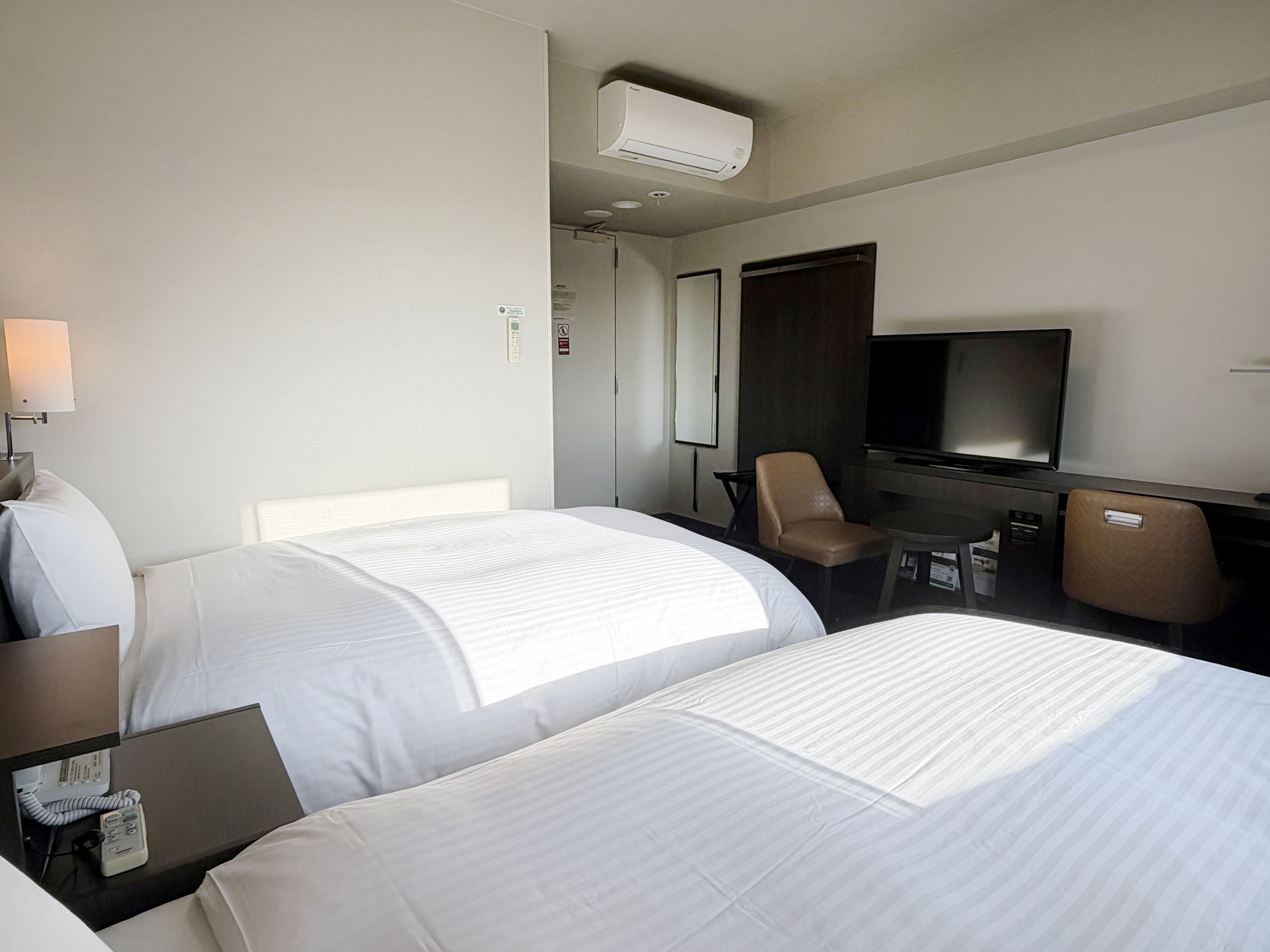 Una camera d'albergo con due letti, una scrivania con telefono, una sedia, una televisione e un condizionatore d'aria.