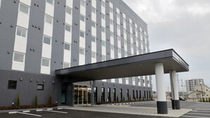 Entrada moderna de hotel com galeria coberta, estacionamento e um edifício alto ao fundo.