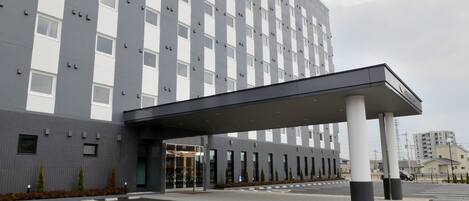 Una entrada de hotel moderna con un paseo techado, un estacionamiento y un edificio alto al fondo.