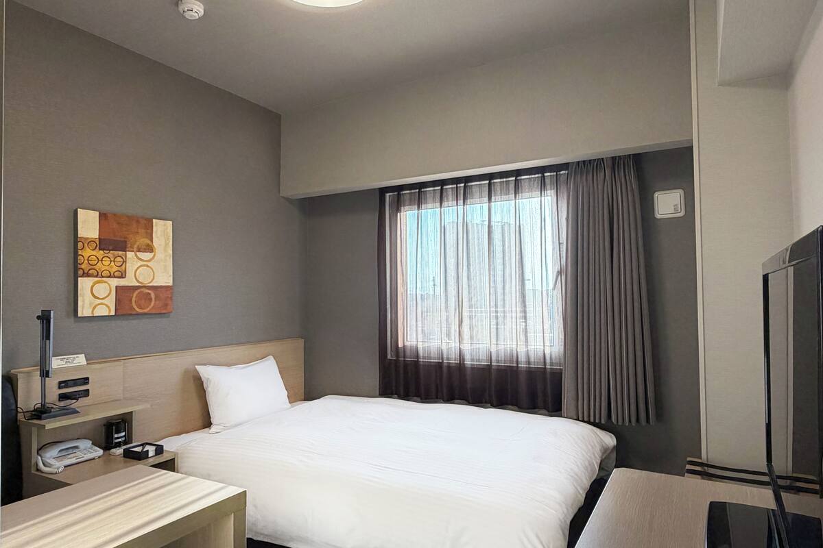 A hotel room with a bed, a desk, a telephone, a lamp, a painting on the wall, and a window with curtains.
