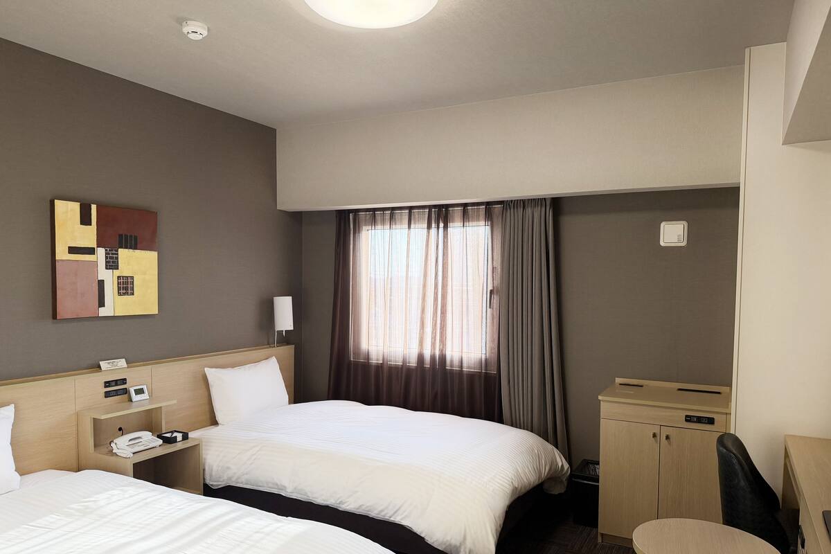 A hotel room with two beds, a desk, a chair, a small table, a telephone, and a wall-mounted light.