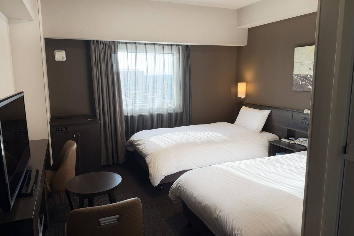 A hotel room with two beds, a desk, a chair, a TV, and a window with curtains.