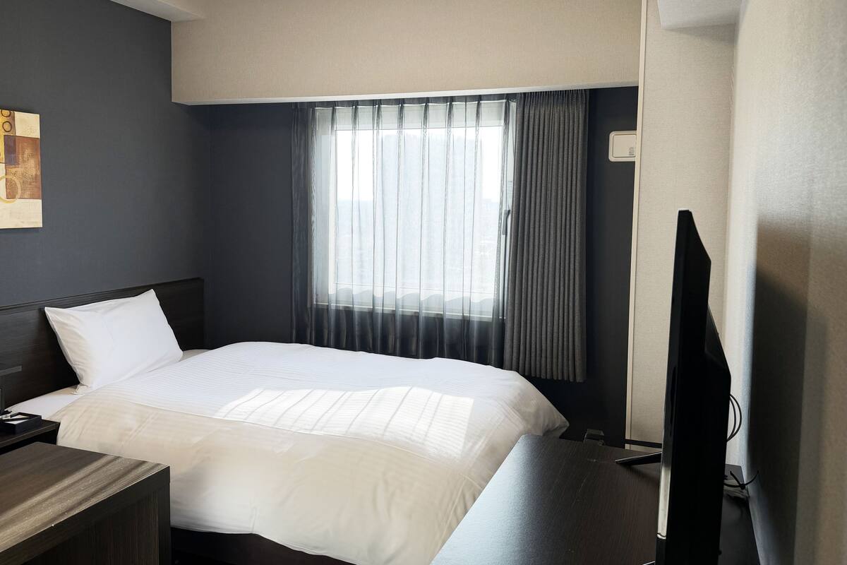 A hotel room with a bed, a bedside table, a TV, and a window with curtains.