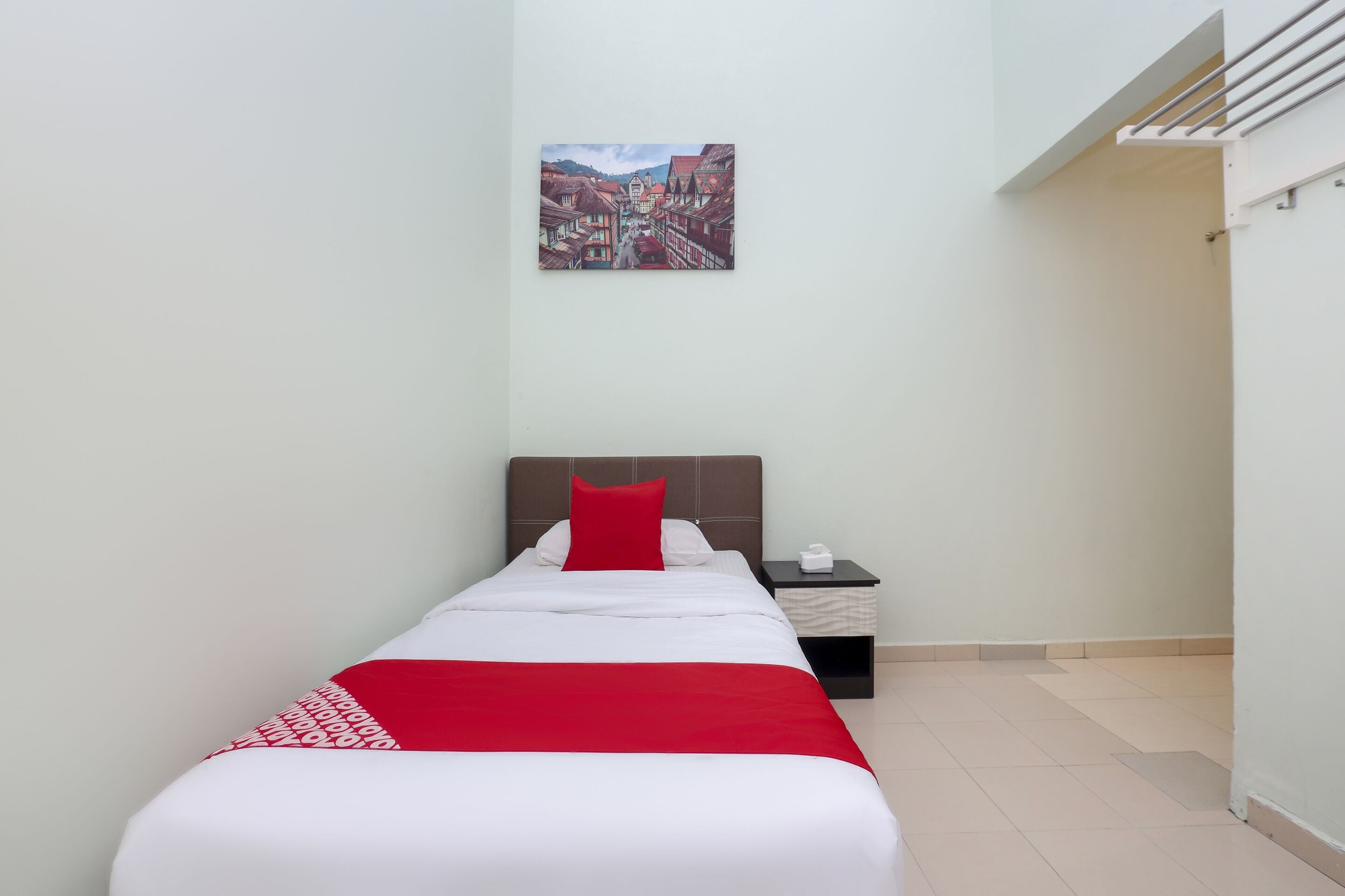 Habitación de hotel con cama, mesita de noche, colcha roja y blanca, y un cuadro con una escena de pueblo en la pared.