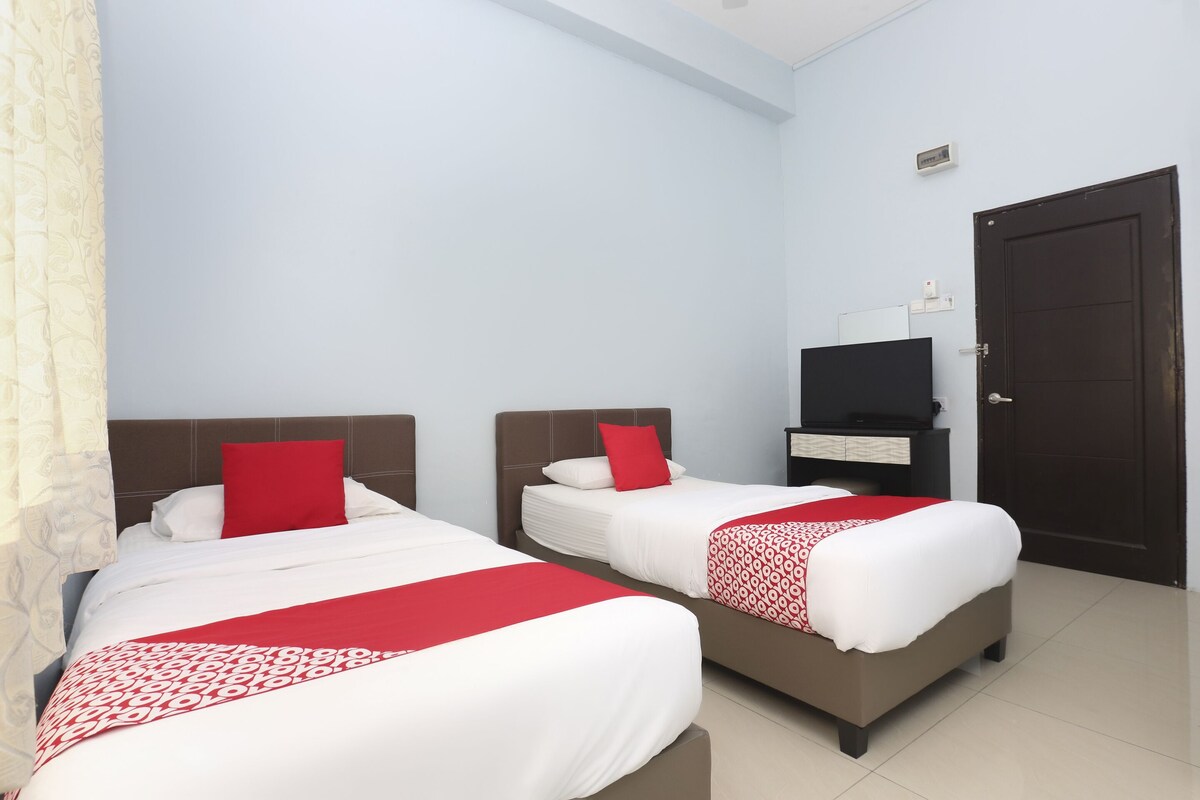 Une chambre d’hôtel avec deux lits, chacun recouvert d’une courtepointe rouge et blanche, doté d’une tête de lit marron et d’un oreiller rouge. Un téléviseur est fixé au mur et une porte se trouve sur le côté droit.